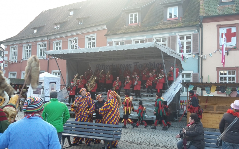 eine Blaskapelle spielt im Schutz der Tribüne. Im Vordergrund Zuschauer und maskierte Narren